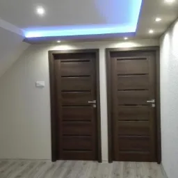 Dwie ciemnobrązowe, nowoczesne drzwi wewnętrzne z poziomymi panelami, zamontowane w pokoju ze skośnym sufitem, oświetlenie LED w suficie podwieszanym.