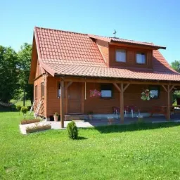 Drewniany dom z czerwonym dachem i zadaszonym tarasem, na którym znajdują się krzesła i stół. Na trawniku widoczny hamak.