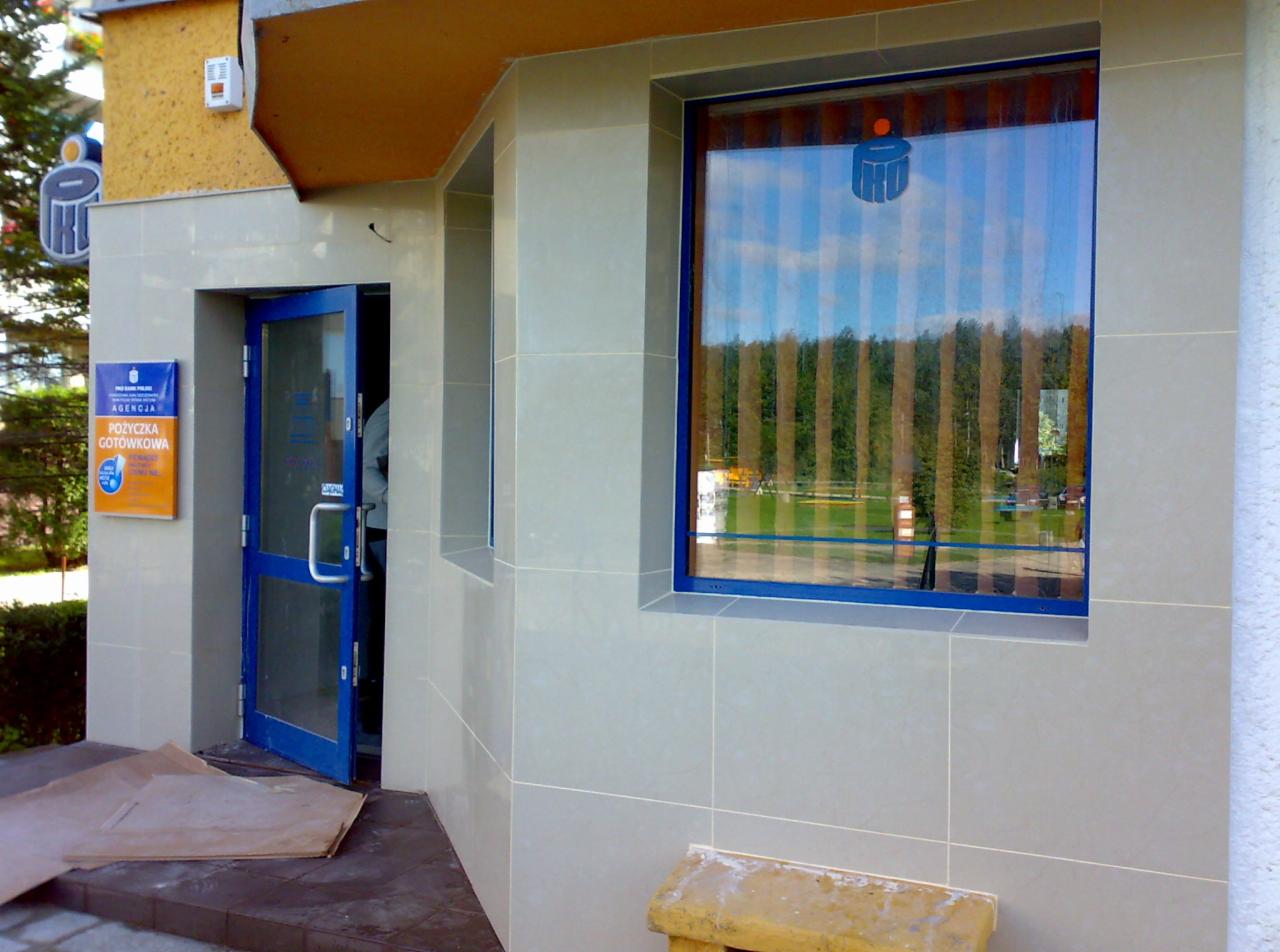 Elewacja budynku wykończona jasnoszarą płytką ceramiczną, z niebieską stolarką okienną i drzwiową, widoczne logo firmy w oknie.