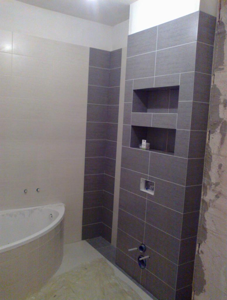 Łazienka w trakcie wykańczania: szare płytki ułożone poziomo z białymi fugami, wnęki w ścianie wyłożone tymi samymi płytkami, fragment wanny, ściana w stanie surowym, widoczne rury doprowadzające...