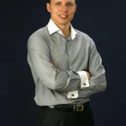 Portret uśmiechniętego mężczyzny w szarej koszuli w paski z białym kołnierzykiem i mankietami, skrzyżowane ramiona na ciemnym tle.