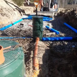 Montaż szamba ekologicznego z rurami drenażowymi na żwirowym podłożu, widoczny wykop i koparka w tle.