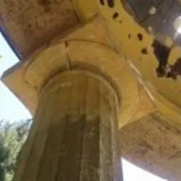 Detal architektoniczny kolumny z widocznymi pęknięciami i odpadającą farbą, skierowany ku górze, z fragmentem niebieskiego nieba i zieleni drzew w tle.