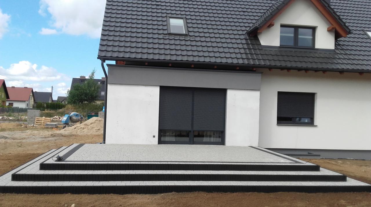 Nowoczesny taras z kostki brukowej w odcieniach szarości, z trzema stopniami obramowanymi ciemną kostką, przylegający do białego domu z szarą stolarką okienną i roletami zewnętrznymi.