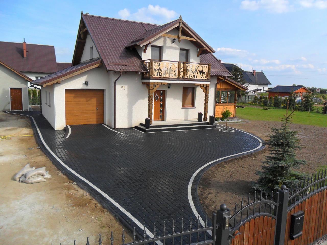 Czarna kostka brukowa z białym obrzeżem prowadząca do domu z drewnianym garażem i balkonem z ażurową balustradą, widok zza metalowego ogrodzenia.