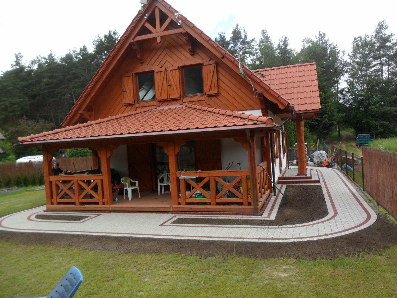 Nowo ułożony chodnik z kostki brukowej w odcieniach szarości i czerwieni, prowadzący do drewnianego domu z werandą, widok z perspektywy trawnika.