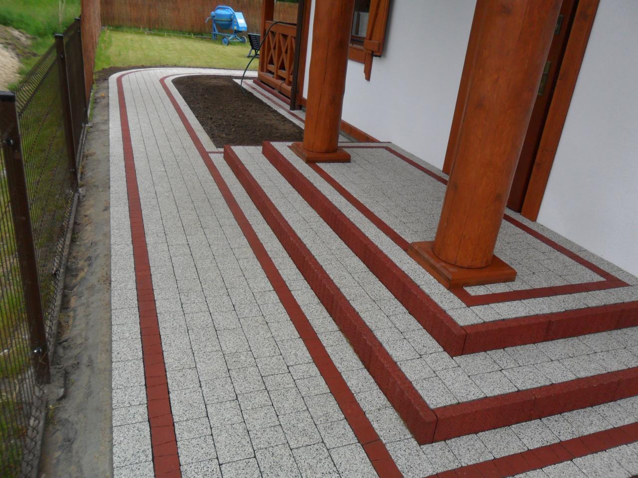 Ułożona szara kostka brukowa z czerwoną obwódką tworząca ścieżkę i schody przed domem z drewnianymi kolumnami, widoczny fragment ogrodzenia i trawnika.