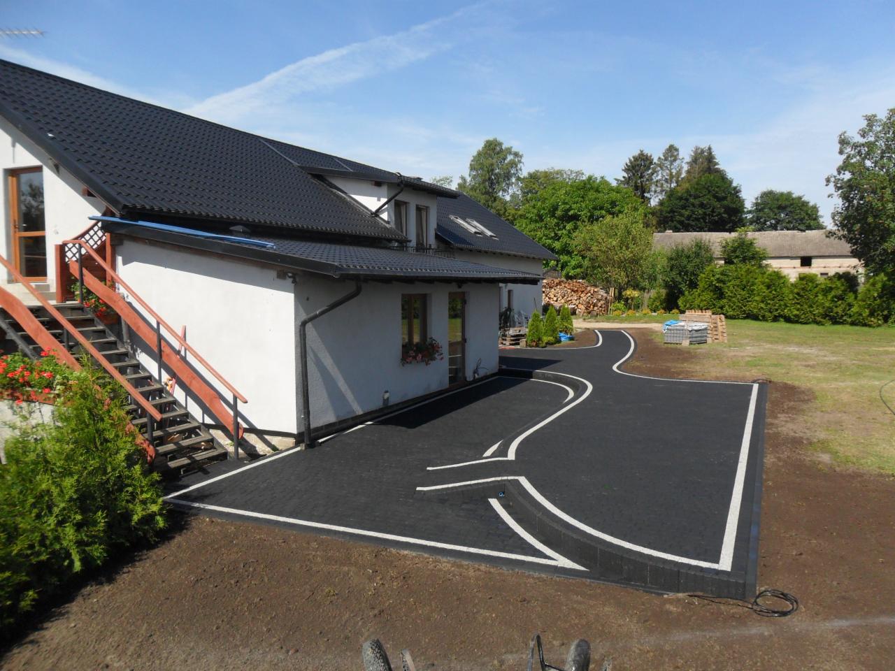 Nowoczesna nawierzchnia z czarnej kostki brukowej z białymi wstawkami, prowadząca od domu z drewnianymi schodami i kwiatami w donicach, przez trawnik w kierunku sterty drewna.