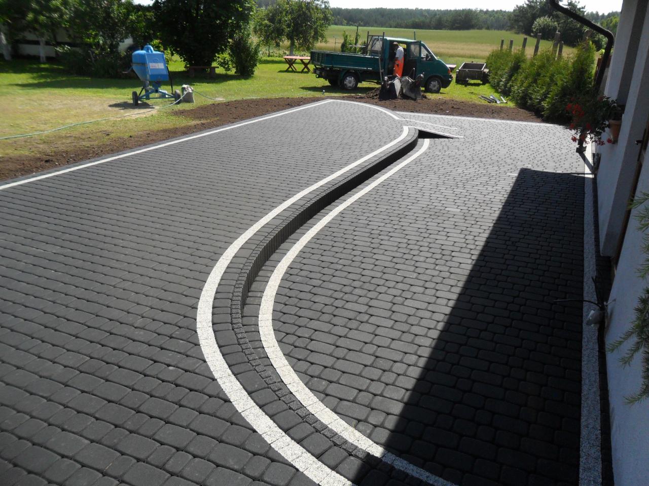 Ukończony podjazd z szarej kostki brukowej z białymi liniami wyznaczającymi łuk, widoczny samochód dostawczy i mieszarka do betonu w tle.