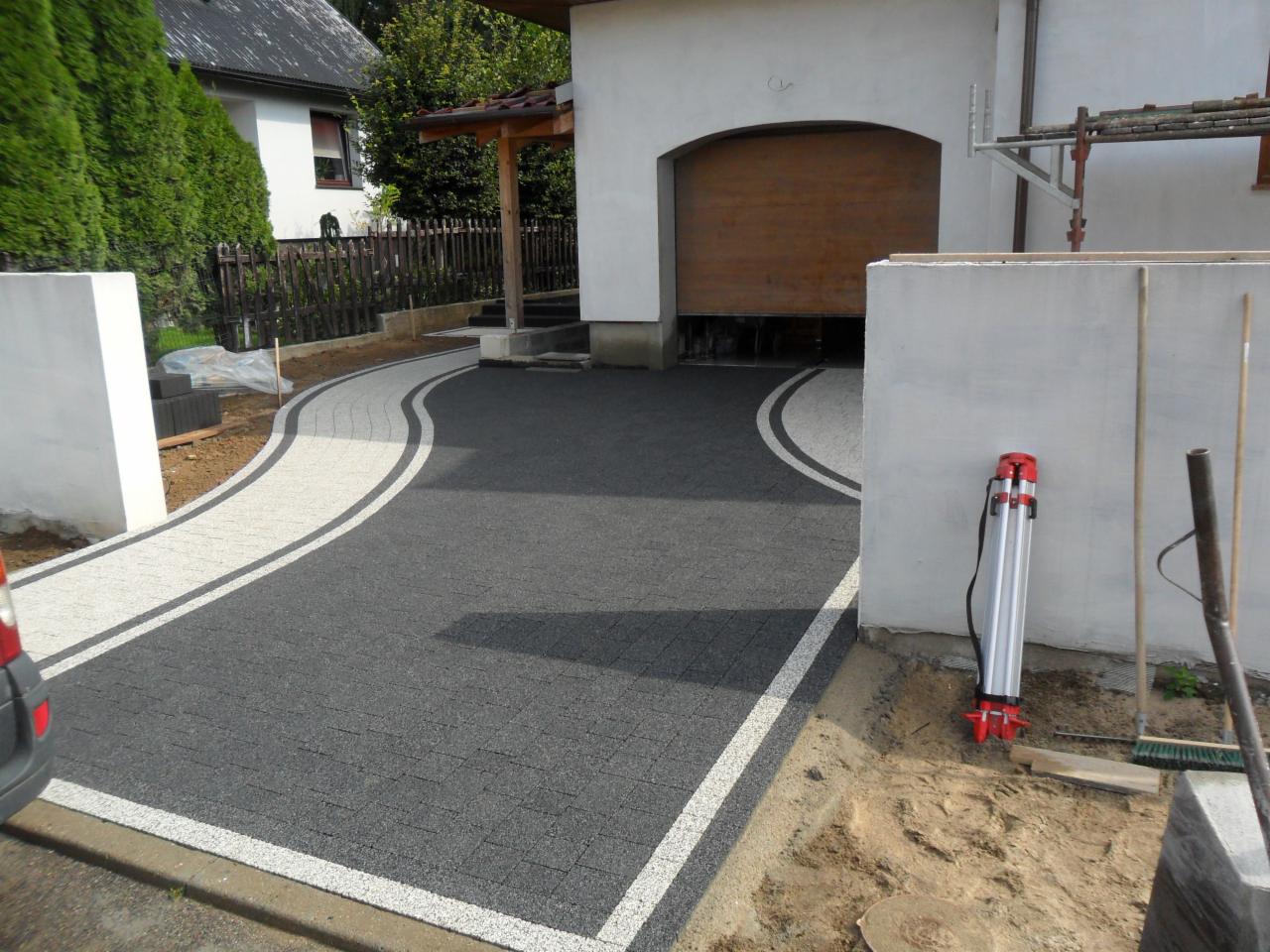 Zakrzywiony podjazd z kostki brukowej w kolorze szarym i białym, prowadzący do garażu z drewnianą bramą, obok biała ściana z niwelatorem.