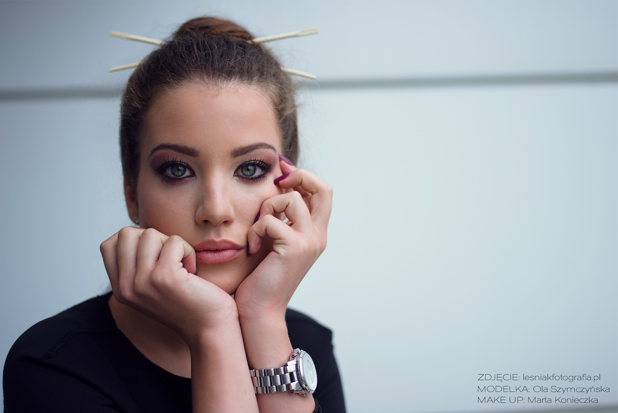 Portret młodej kobiety z upiętymi włosami, w których umieszczono pałeczki, z intensywnym makijażem oczu i zegarkiem na ręce, opierającej twarz na dłoniach.