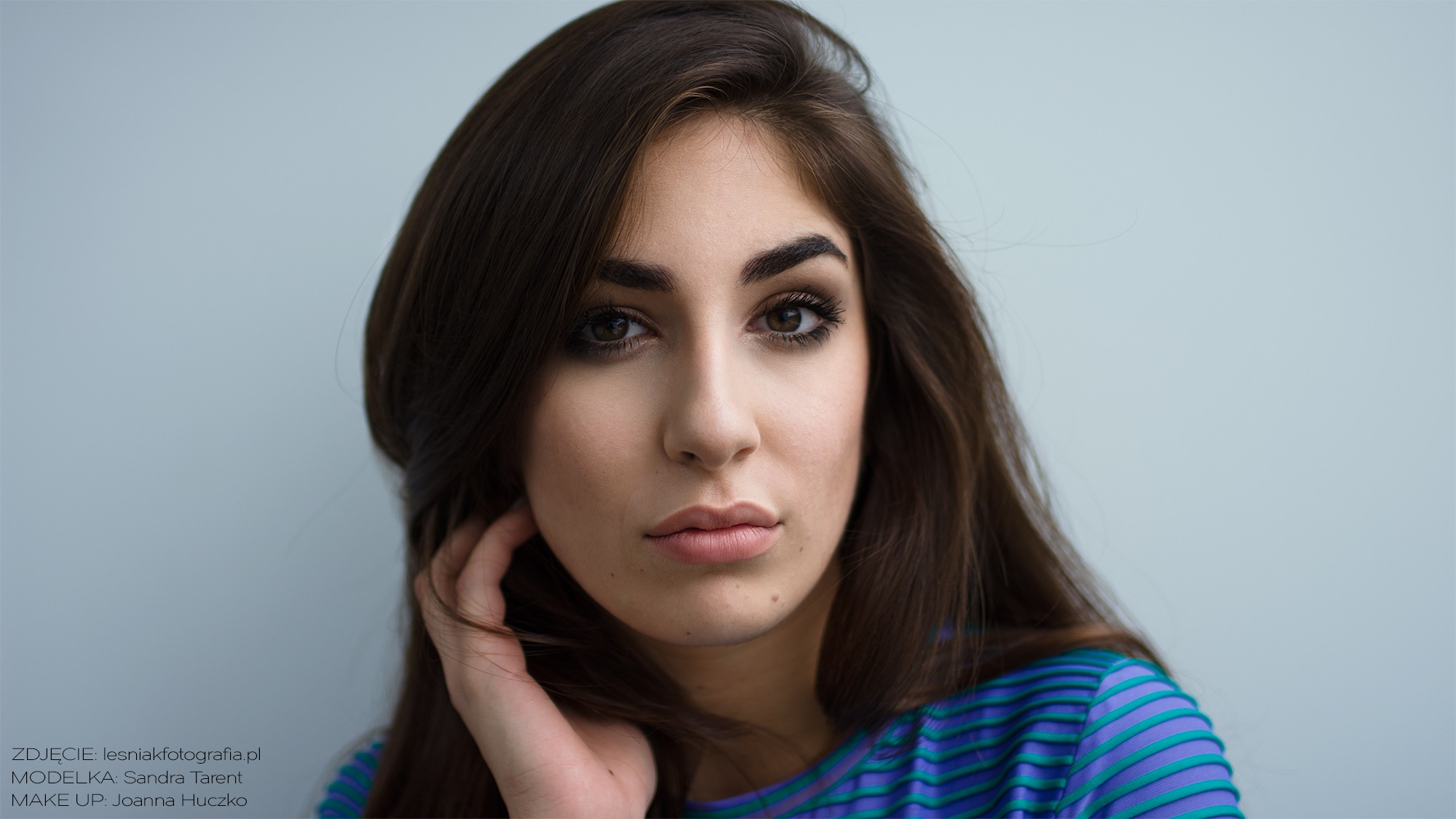 Portret kobiety z brązowymi włosami i intensywnym makijażem oczu, w bluzce w paski, trzymającej dłoń przy twarzy.