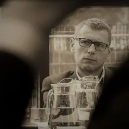 Portret biznesowy mężczyzny w okularach, zamyślony, w tle dzbanki z wodą, stonowana kolorystyka.