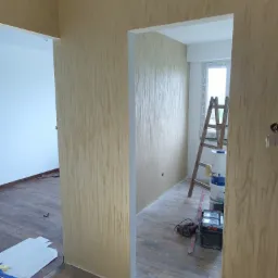 Ściana z dekoracyjną fakturą w odcieniach beżu, widoczne narzędzia malarskie, drabina i pojemniki z farbą, w tle okno w trakcie remontu.