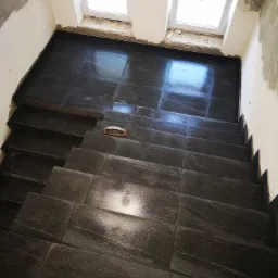 Schody wyłożone ciemnymi, połyskującymi płytkami w trakcie prac w Alwerni, widoczne dwa okna dachowe i ślady po pracach wykończeniowych na ścianach, na jednym ze stopni leży kielnia.