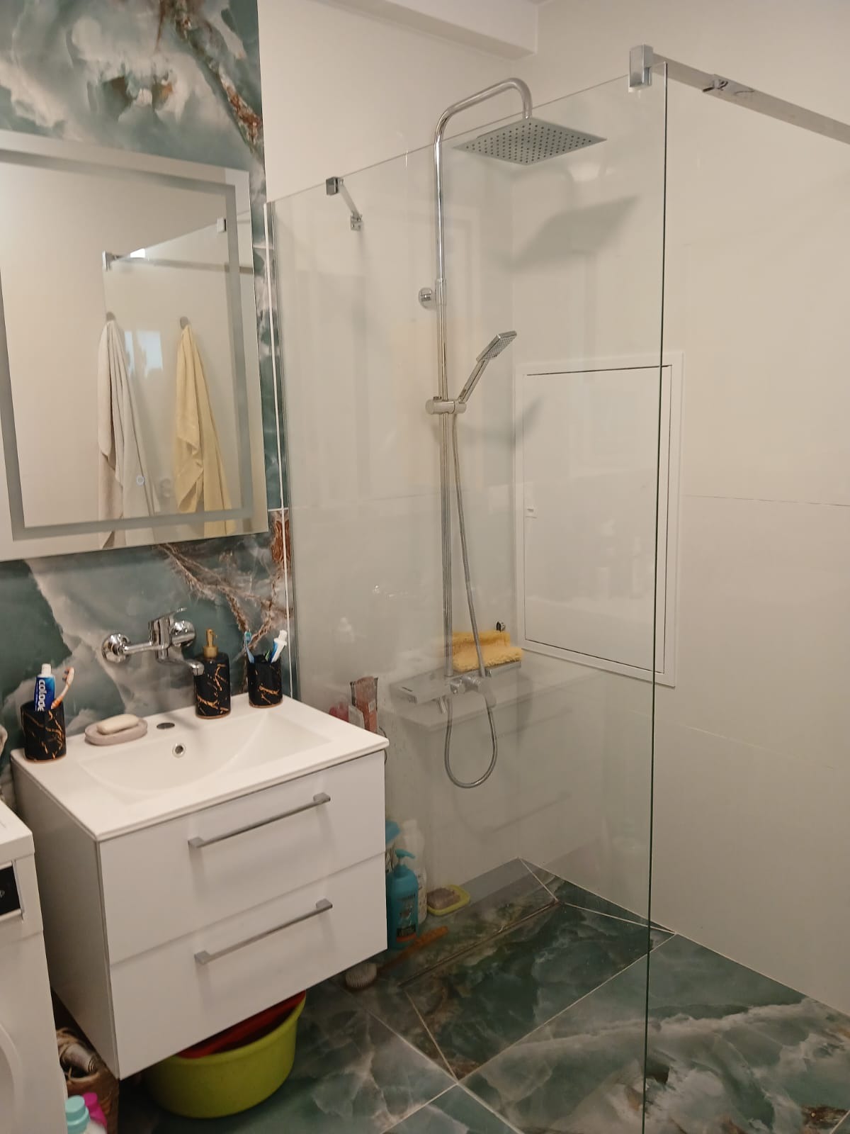 Nowoczesna łazienka po remoncie: biała umywalka z szafką, szklana kabina prysznicowa typu walk-in, zielona marmurowa podłoga i dekoracyjne lustro z podświetleniem.