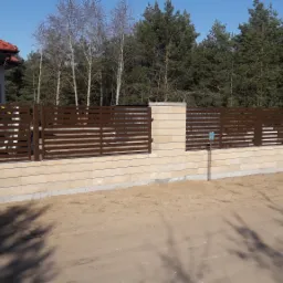 Nowoczesne ogrodzenie z poziomych, brązowych paneli zamontowanych na słupkach z beżowego kamienia, z widocznym fragmentem żółtego samochodu dostawczego i domu w tle.