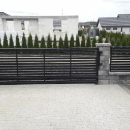 Nowoczesna brama wjazdowa z poziomych profili stalowych, osadzona na szarych, betonowych słupkach, z kostką brukową na podjeździe, w tle budynek w stanie surowym otoczony tujami.