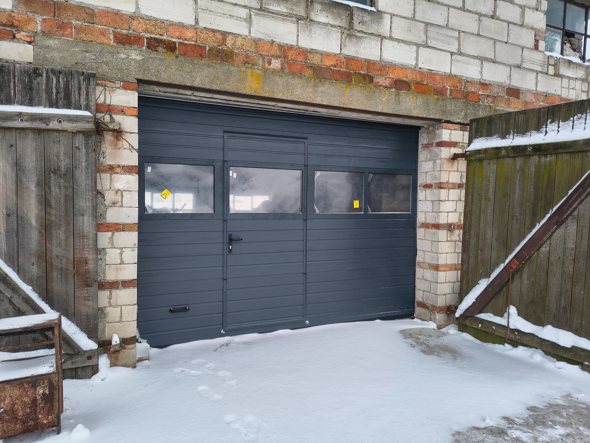 Grafitowa brama garażowa segmentowa z oknami, zamontowana w murowanym garażu z drewnianymi drzwiami obok, pokryta śniegiem. Widoczne ślady na śniegu.