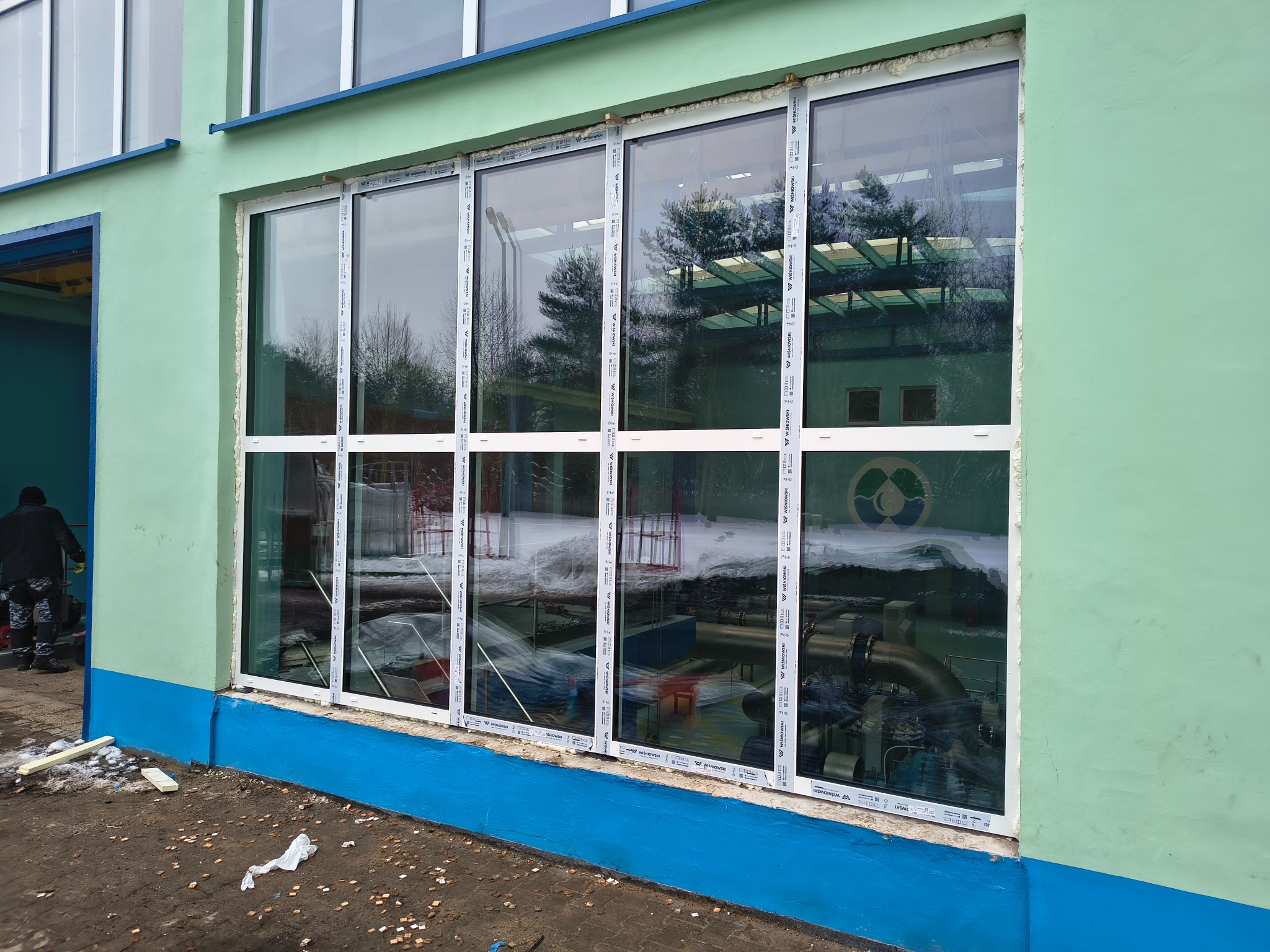 Montaż okien panoramicznych w budynku o zielonej elewacji. Widoczne białe ramy okienne i odbicie wnętrza budynku w szybach. Prace montażowe w toku, widoczny pracownik.
