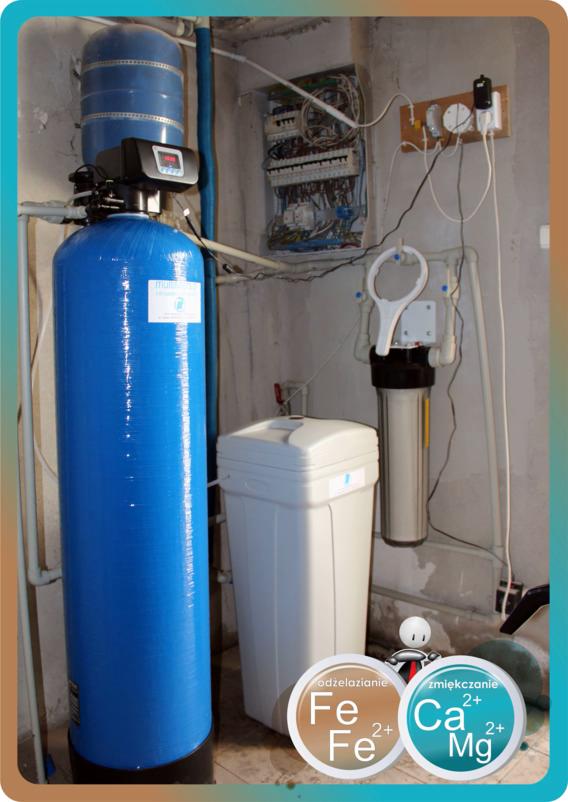 Zestaw filtrów do uzdatniania wody w pomieszczeniu technicznym, widoczny duży niebieski filtr, biały zbiornik na sól, filtr narurowy oraz skrzynka elektryczna na ścianie.