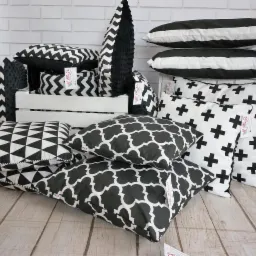 Kolekcja dekoracyjnych poduszek w czarno-białe wzory, ułożonych na drewnianej podłodze przed białą ceglaną ścianą, niektóre w białej drewnianej skrzynce.