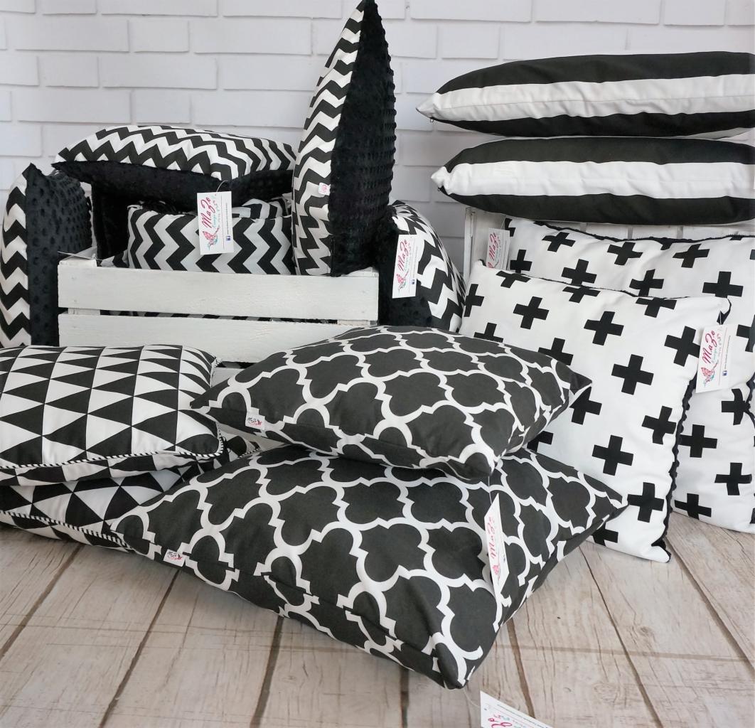 Kolekcja dekoracyjnych poduszek w czarno-białe wzory, ułożonych na drewnianej podłodze przed białą ceglaną ścianą, niektóre w białej drewnianej skrzynce.