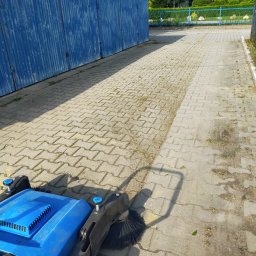 Firma Sprzątająca - Niebieska zamiatarka do kostki brukowej na tle metalowego garażu i fragmentu czystej oraz zabrudzonej kostki brukowej.