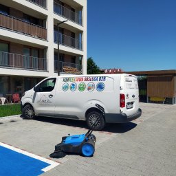 Firma Sprzątająca - Biały samochód dostawczy z logo firmy sprzątającej na parkingu z kostki brukowej, obok stoi niebieska zamiatarka do kostki.