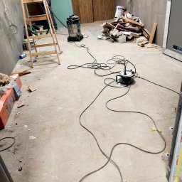 Firma Sprzątająca - Wnętrze w trakcie remontu: drabina, odkurzacz przemysłowy, gruz i resztki materiałów budowlanych na podłodze i przy ścianach, widoczne kable elektryczne.