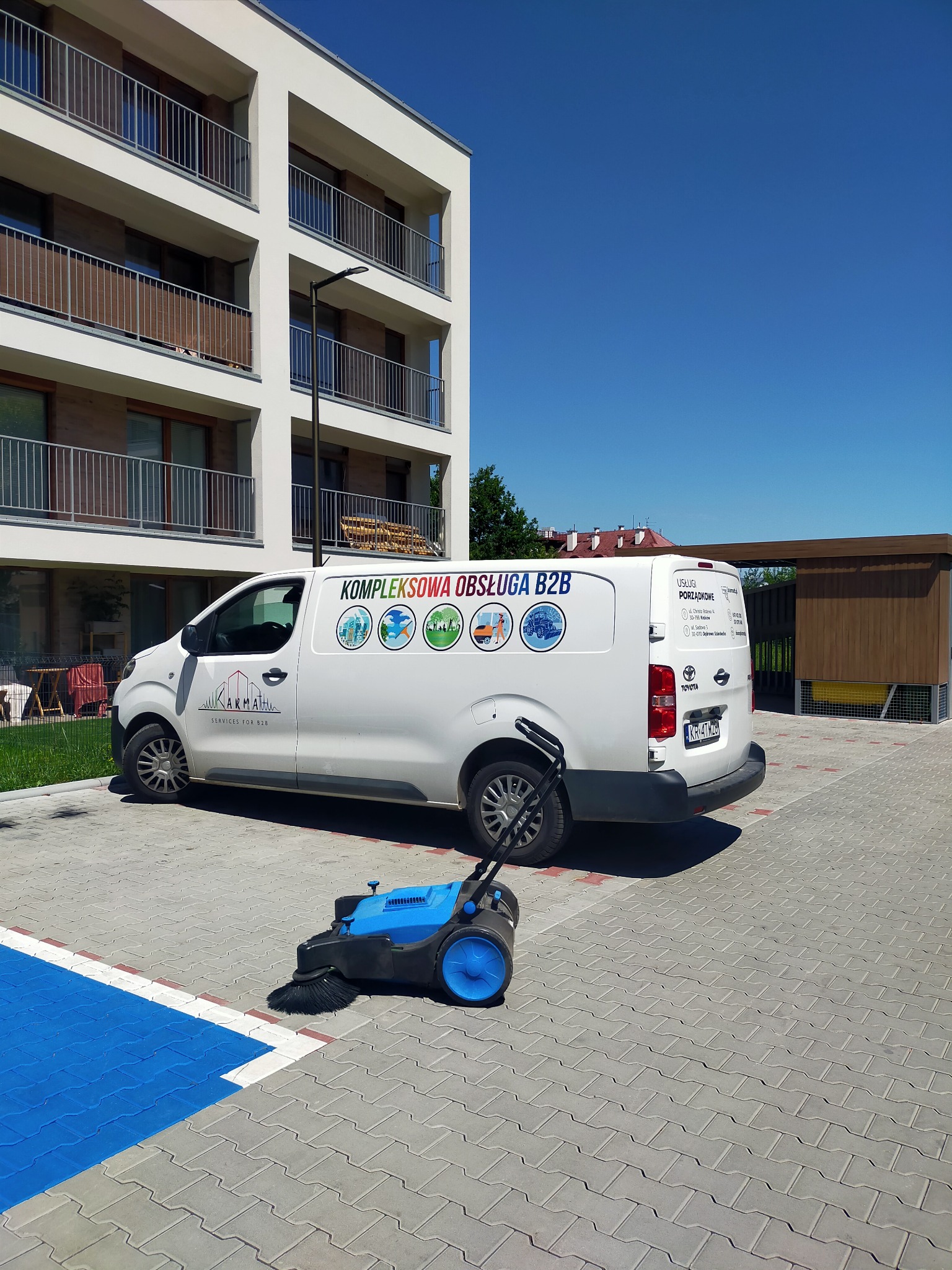 Biały samochód dostawczy z logo firmy sprzątającej na parkingu z kostki brukowej, obok stoi niebieska zamiatarka do kostki.