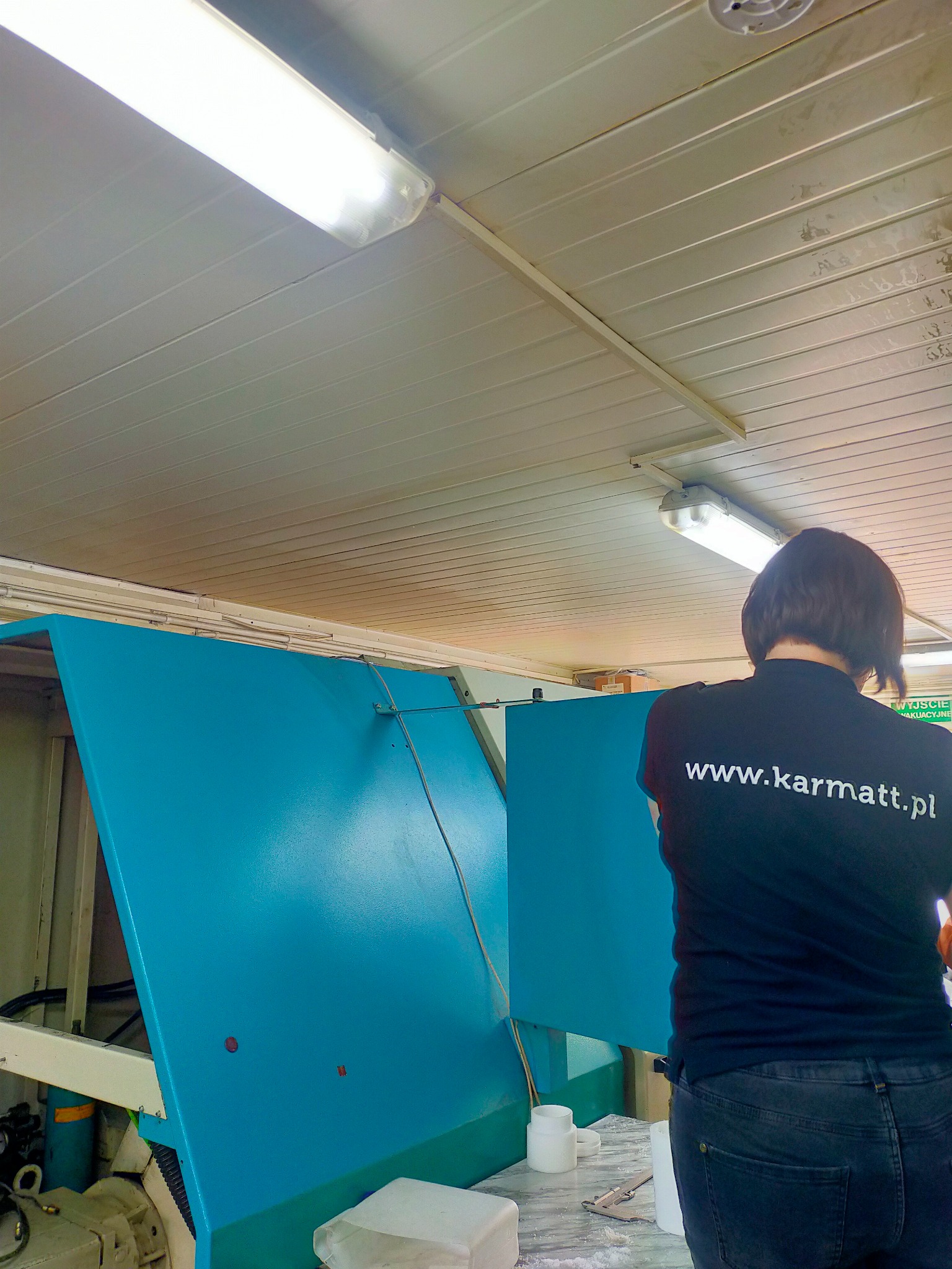 Pracownica w czarnej koszulce z adresem www.karmatt.pl obsługuje niebieską maszynę przemysłową w hali produkcyjnej pod oświetleniem jarzeniowym.