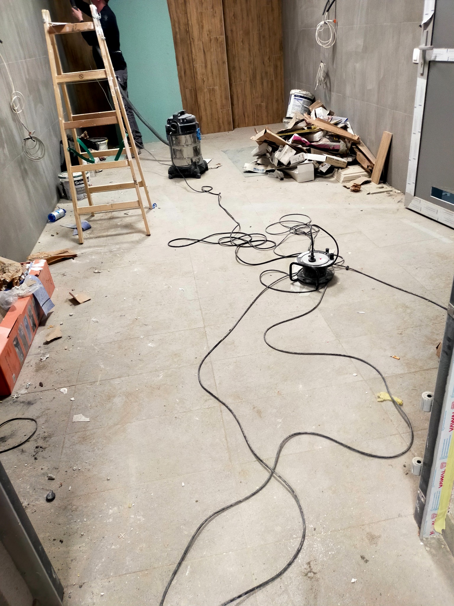 Wnętrze w trakcie remontu: drabina, odkurzacz przemysłowy, gruz i resztki materiałów budowlanych na podłodze i przy ścianach, widoczne kable elektryczne.