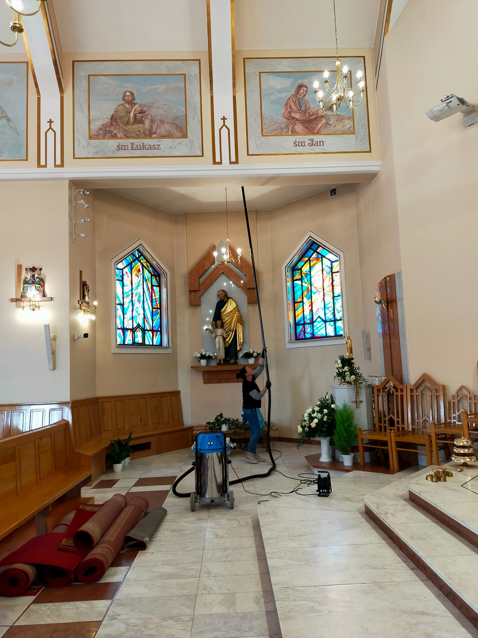 Pracownica firmy sprzątającej używa odkurzacza teleskopowego do czyszczenia wysokich ścian w kościele, widoczne witraże i obrazy religijne, na podłodze zwinięte dywany i profesjonalny odkurzacz...