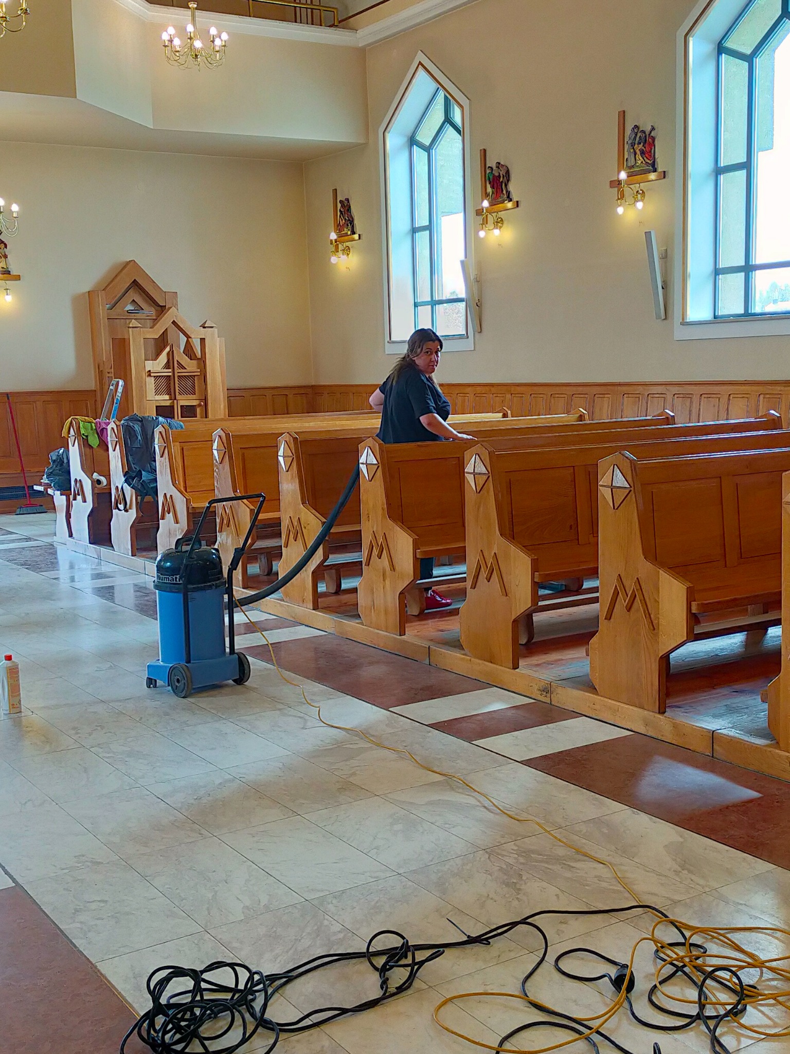Kobieta w trakcie porządkowania wnętrza kościoła, używa odkurzacza przemysłowego Numatic do czyszczenia podłogi pomiędzy drewnianymi ławkami.
