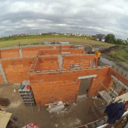 Wczesny etap budowy domu jednorodzinnego: widok z góry na wznoszone ściany z czerwonej cegły, widoczny pracownik w pomarańczowej kamizelce, teren budowy z żółtą koparką w tle i zabudowaniami...