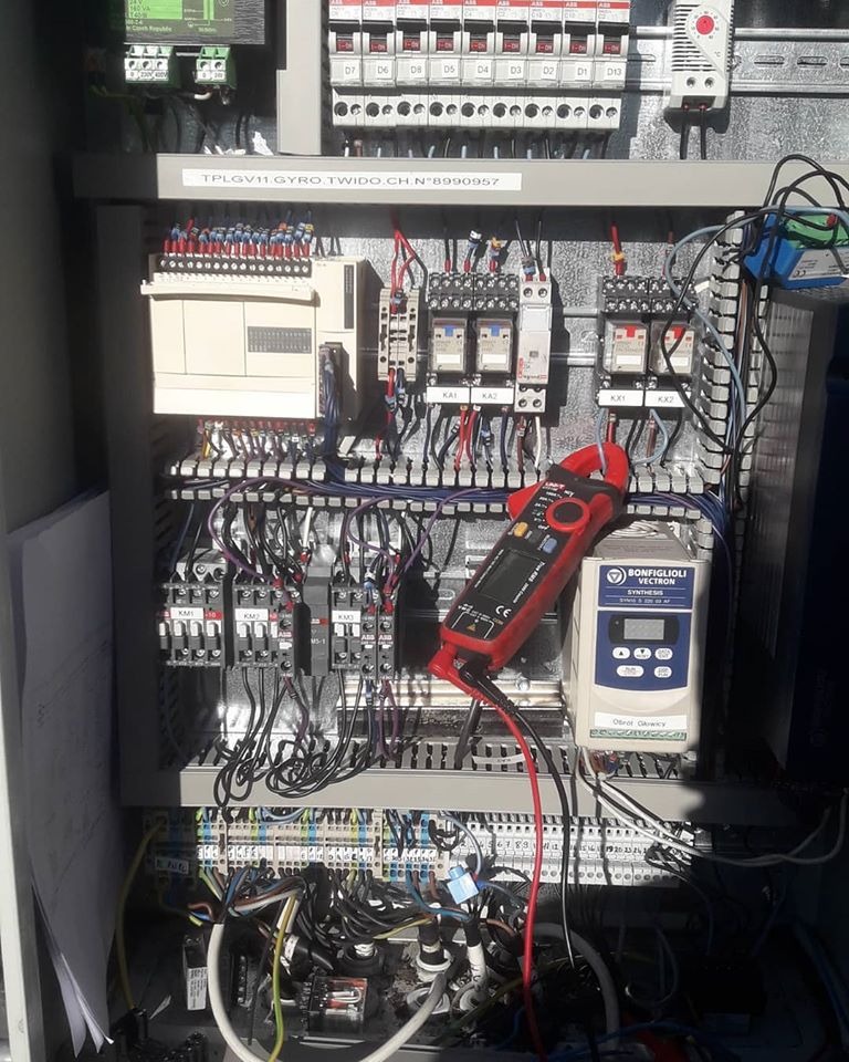 Pomiar parametrów elektrycznych w szafie sterowniczej za pomocą miernika cęgowego, widoczny falownik Bonfiglioli Vectron i liczne przekaźniki.