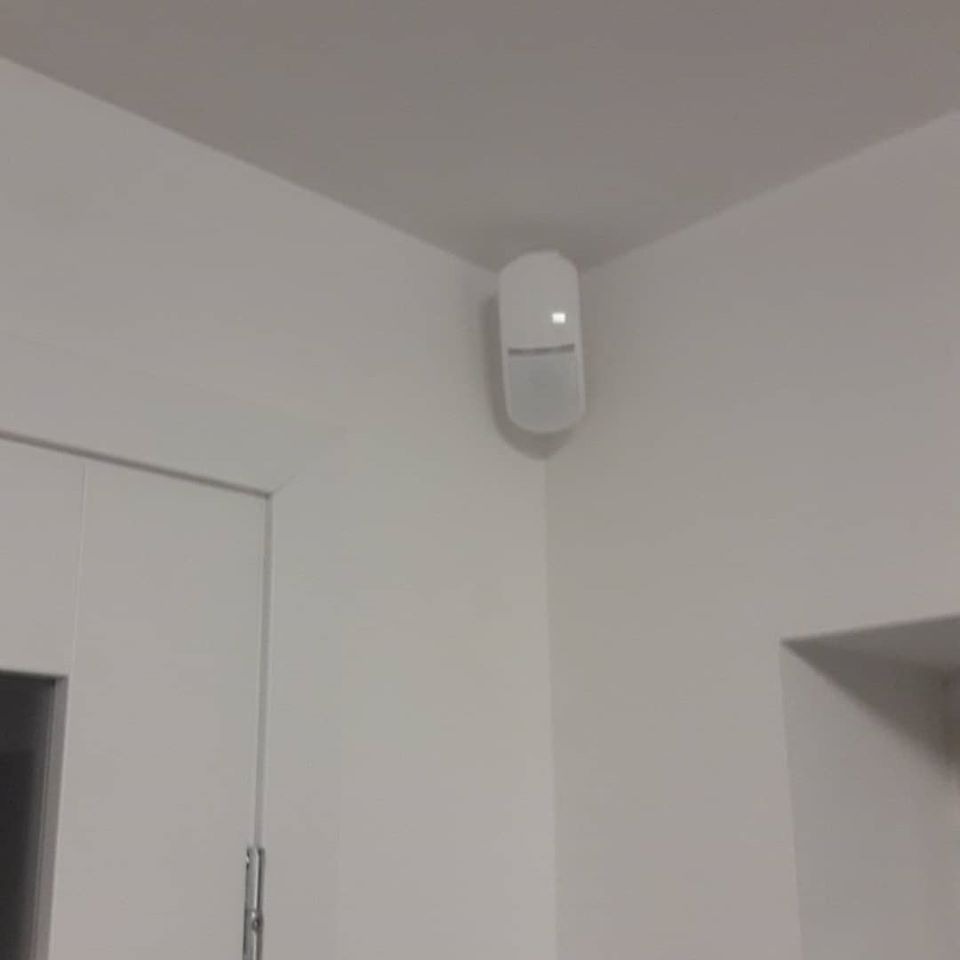 Biały czujnik ruchu zamontowany w rogu pomieszczenia, widoczna biała ściana i fragment futryny drzwi.