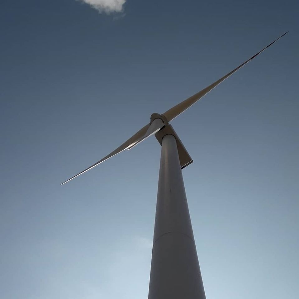 Wysoka, biała turbina wiatrowa sfotografowana z dołu na tle błękitnego nieba z pojedynczą chmurą.