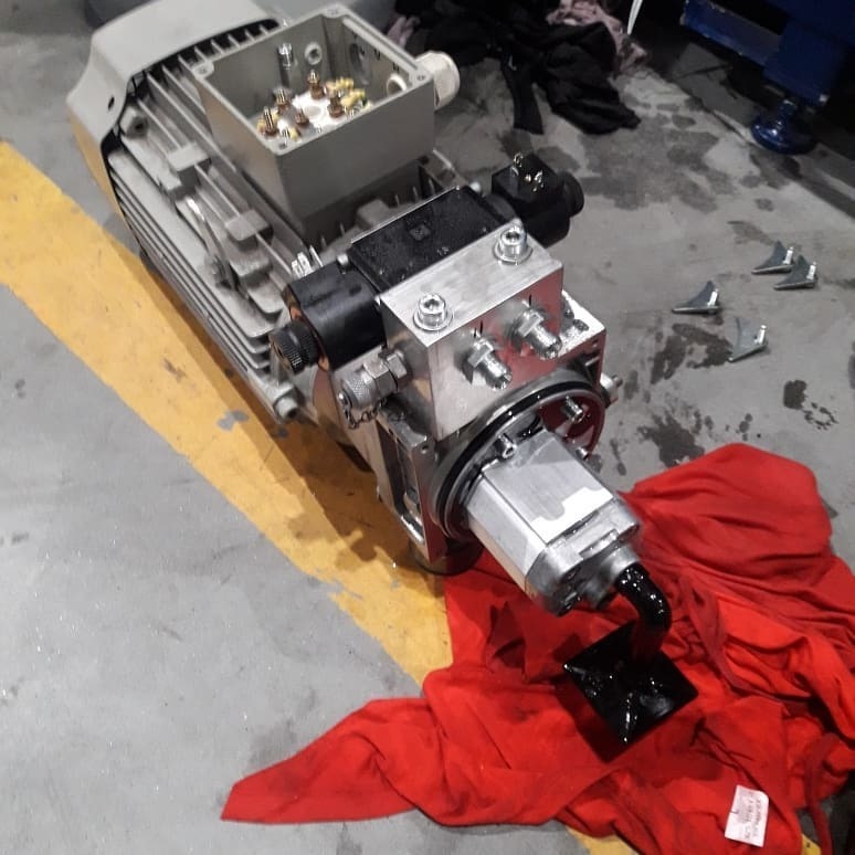 Szary silnik elektryczny z podłączoną pompą hydrauliczną na betonowej podłodze z żółtą linią, widoczne ślady oleju na czerwonej szmacie i podłodze, otwarte złącze elektryczne.