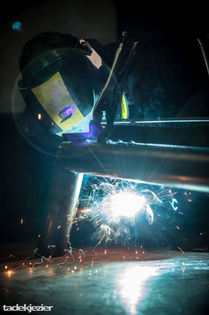 Spawanie metalowej konstrukcji przez spawacza w masce ochronnej, widoczne iskry i dym, ciemne tło.