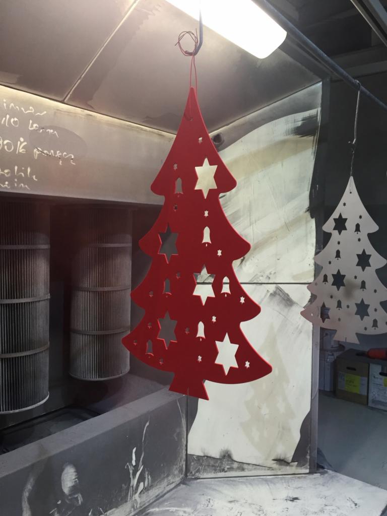Czerwony, metalowy, wycinany laserowo kształt choinki z gwiazdami i dzwoneczkami, wiszący w przemysłowej lakierni proszkowej, obok podobny, biały element.