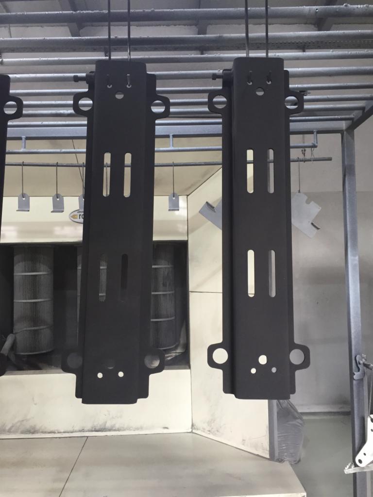 Czarne, metalowe elementy z otworami zawieszone na hakach w lakierni proszkowej, widoczny piec do wypalania farby.