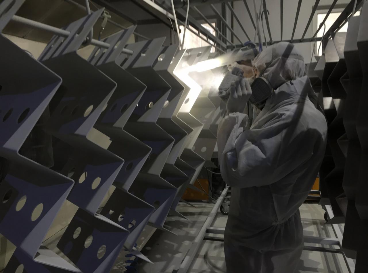 Pracownik w kombinezonie ochronnym maluje proszkowo metalowe elementy o zygzakowatym kształcie, zawieszone na metalowym stelażu.