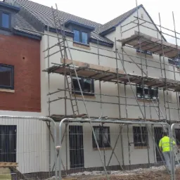 Nowo wybudowany budynek z fragmentem elewacji z czerwonej cegły i otynkowaną, jasną elewacją w trakcie prac wykończeniowych, z rusztowaniami i pracownikiem w odblaskowej kamizelce na placu budowy.