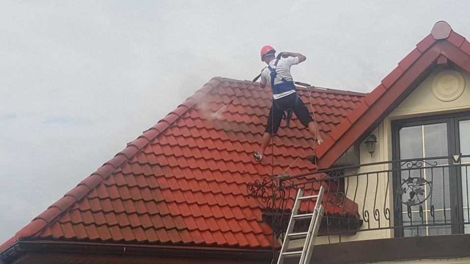 Pracownik w kasku i uprzęży zabezpieczającej myje pod ciśnieniem dach z czerwonej dachówki, widoczna drabina oparta o balkon z ozdobną balustradą.