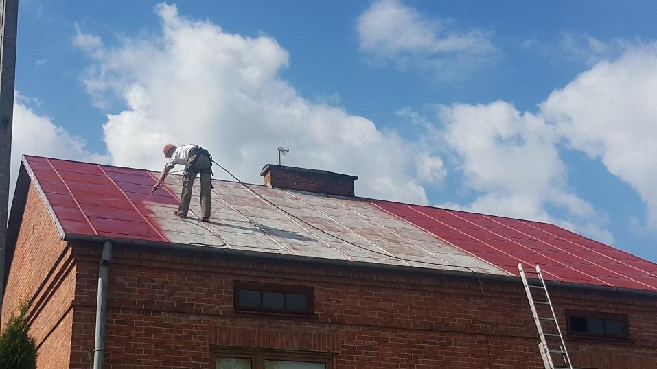 Malowanie dachu z czerwonej blachy falistej przez pracownika stojącego na dachu, widoczna drabina oparta o ceglany budynek, błękitne niebo z chmurami w tle.