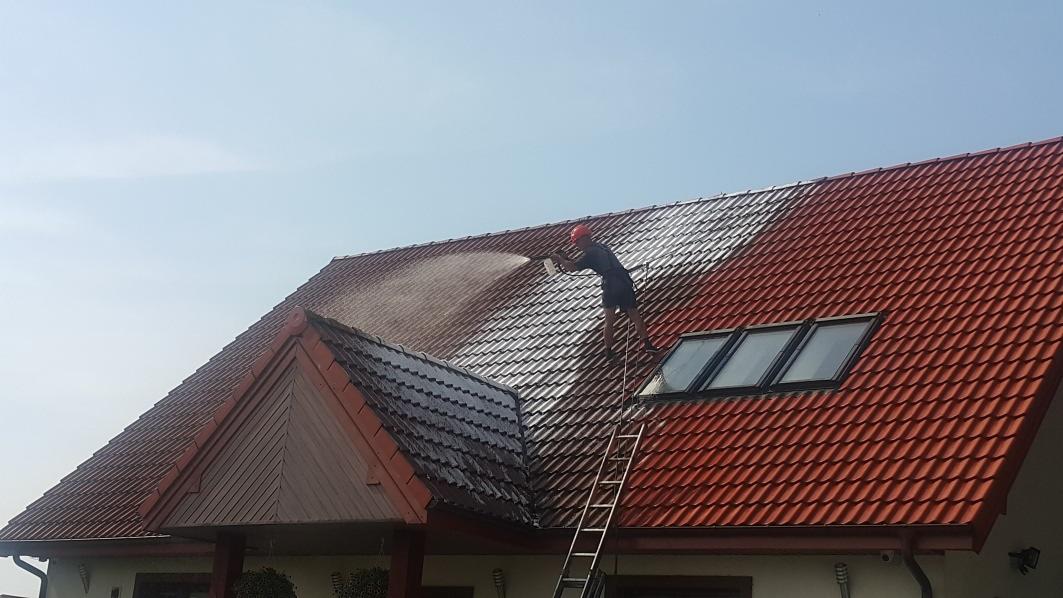 Mężczyzna w kasku i krótkich spodenkach myje dach z czerwonej dachówki za pomocą myjki ciśnieniowej, stojąc na drabinie; część dachu jest już umyta, ukazując ciemniejszy kolor.