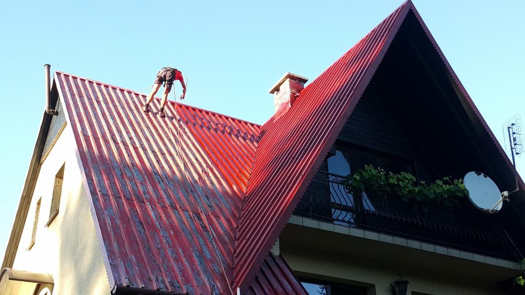 Pracownik zabezpieczony linami, w trakcie pracy na dachu z blachy trapezowej, widoczny fragment budynku z oknami i balkonem.