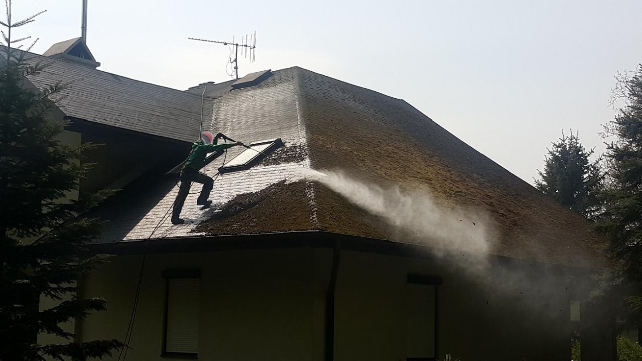 Pracownik w zielonym kombinezonie czyści dach budynku w Wojciechowie za pomocą myjki ciśnieniowej, strumień wody usuwa zabrudzenia z powierzchni dachu.